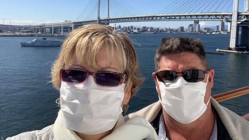 A man and a woman wearing sunglasses and face masks pose in front of a bridge.