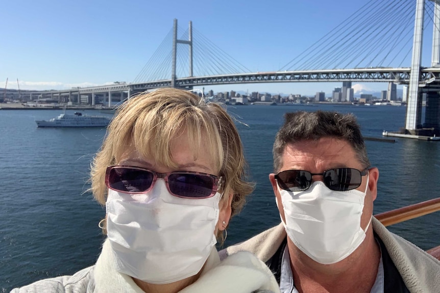 A man and a woman wearing sunglasses and face masks pose in front of a bridge.