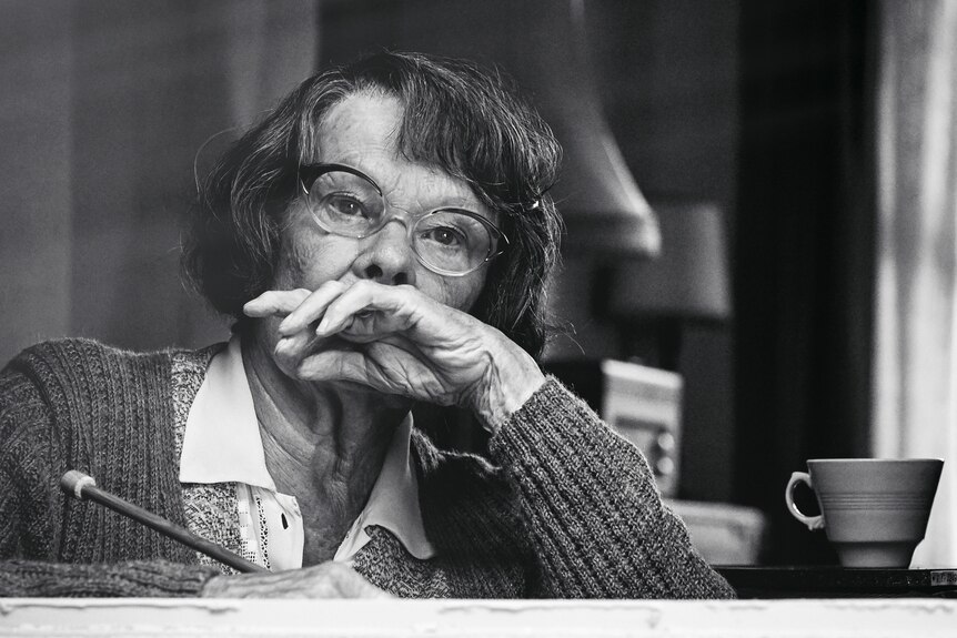 A black and white image of an older woman in glasses looking pensive, her hand to her face, and her other holding a pencil