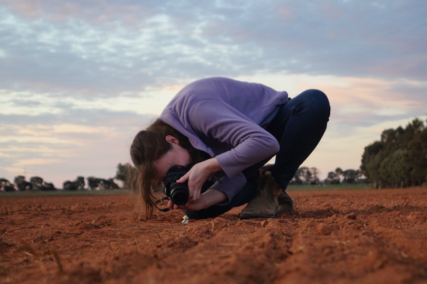Photograph of a young woman in a ploughed field, crouching down to photograph a tiny farmer lego man on the ground.