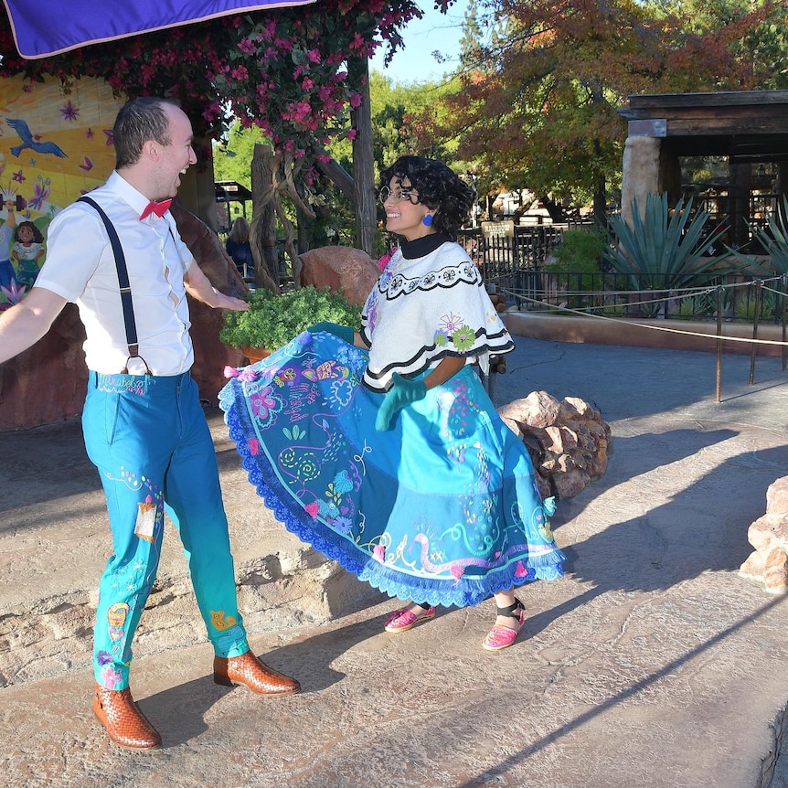 Joel Callen wearing blue trousers dances with a woman dressed in the embroidered blue skirt of Mirabel from Encanto.