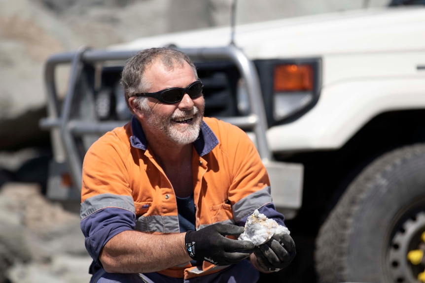 A man wearing high-vis workwear and sunglasses holding a gold rock specimen. 