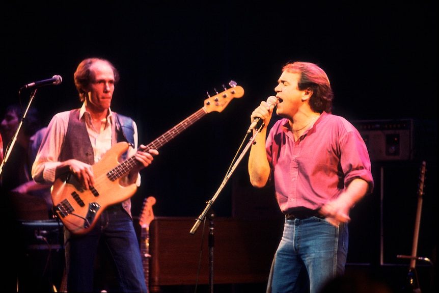 On stage, one man plays bass guitar and the other hands a microphone, yelling into it. They wear tight jeans and flowing tops.