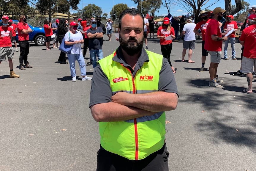 A man with his arms crossed wearing a high-vis vest stands in front of a crowd of people wearing red t-shirts