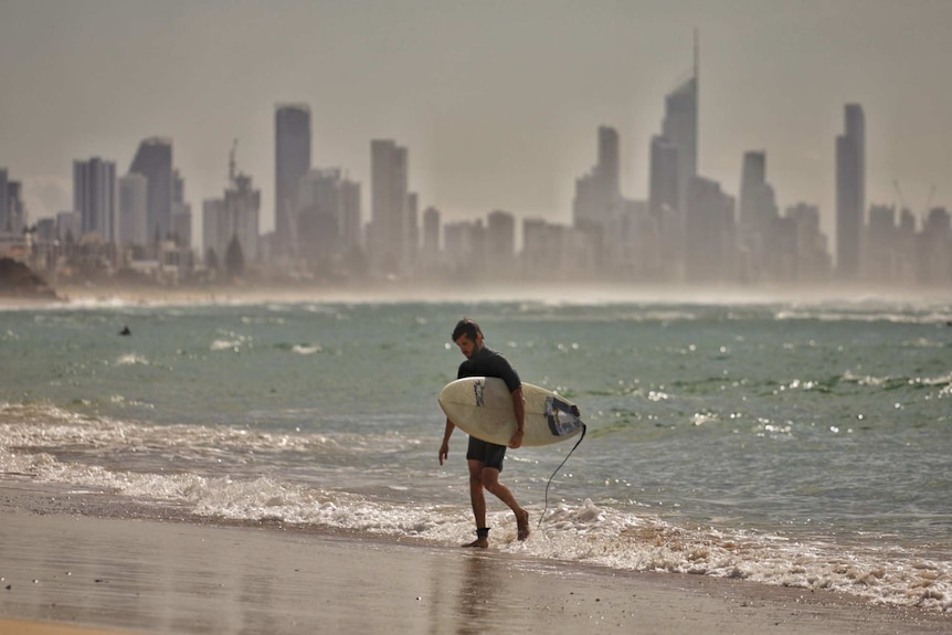 A man exits the surf, holding his board, with skyscrapers in the background.