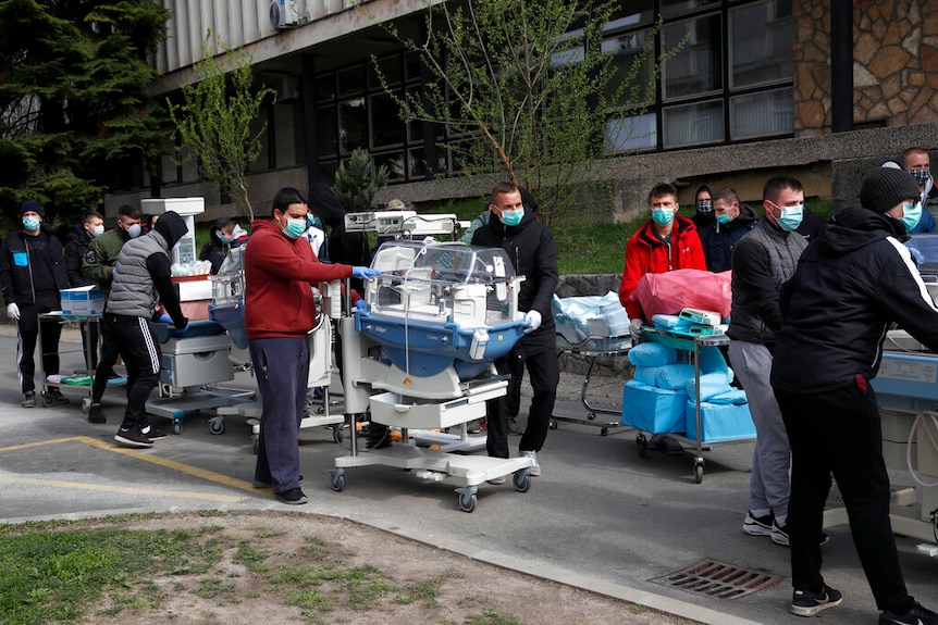 A line of men wearing surgical masks wheel infant incubators in a line outside a grey hospital building.