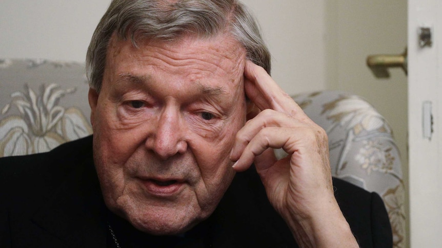 A man wearing a black robe holds his fingers to his temple as he speaks.