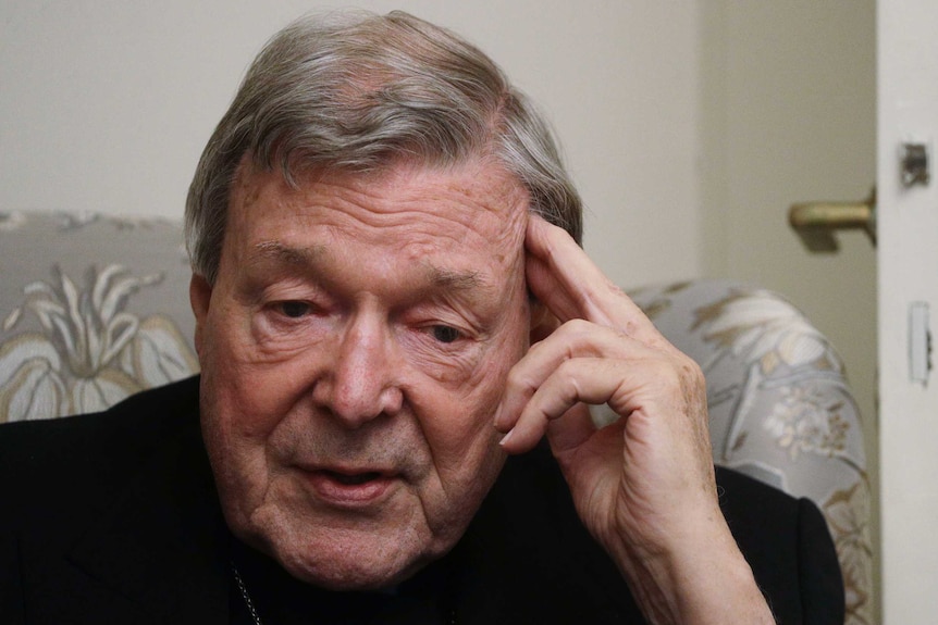 A man wearing a black robe holds his fingers to his temple as he speaks.