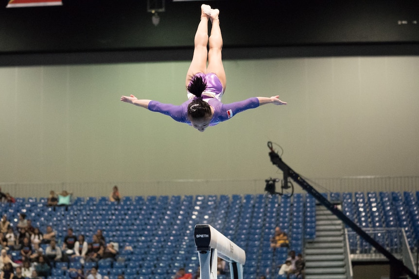 A woman with brown ponytail and purple leotard lands horizontally, with head facing down, from a very high flip through the air.