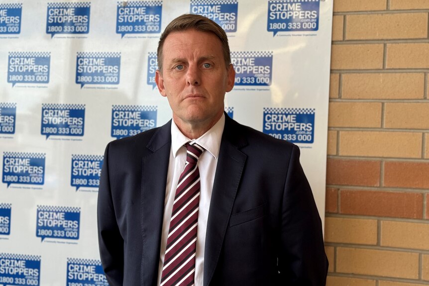 A stern-looking man in a dark suit stands in front of a Crime Stoppers backdrop.