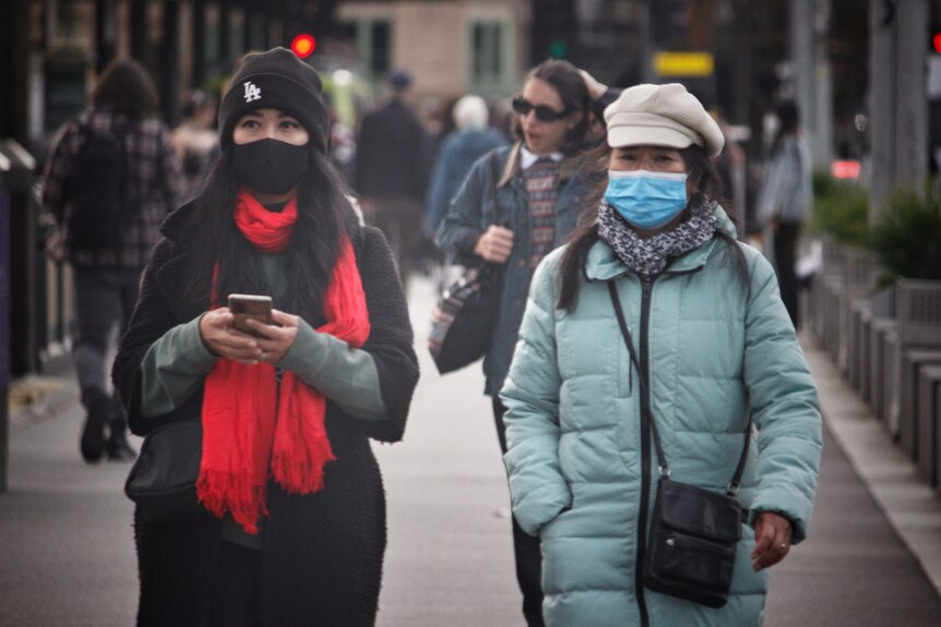 Two women in surgical masks and warm clothing walk down a CBD street.