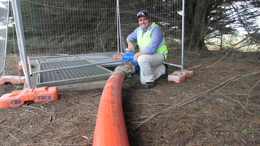 A man in a hi-vis vest kneels near a large orange pipe, which is connected to a water main in rural Victoria.