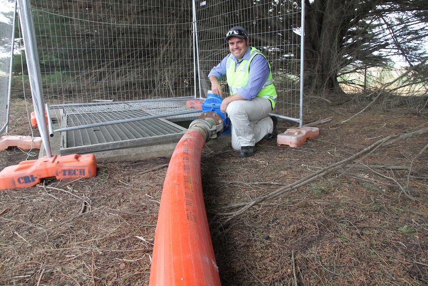 A man in a hi-vis vest kneels near a large orange pipe, which is connected to a water main in rural Victoria.