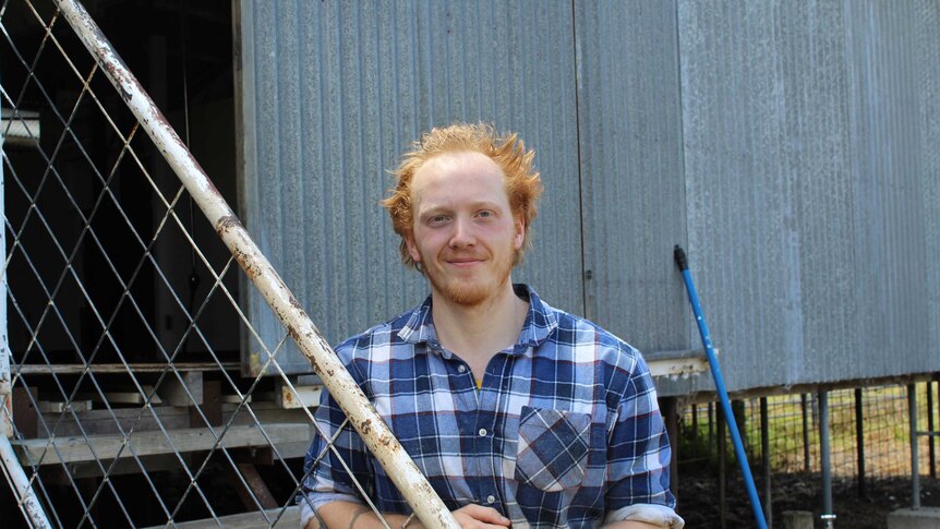 A red-haired man in a blue and white checked shirt leans on a stair-railing in front of a corrugated iron shed.