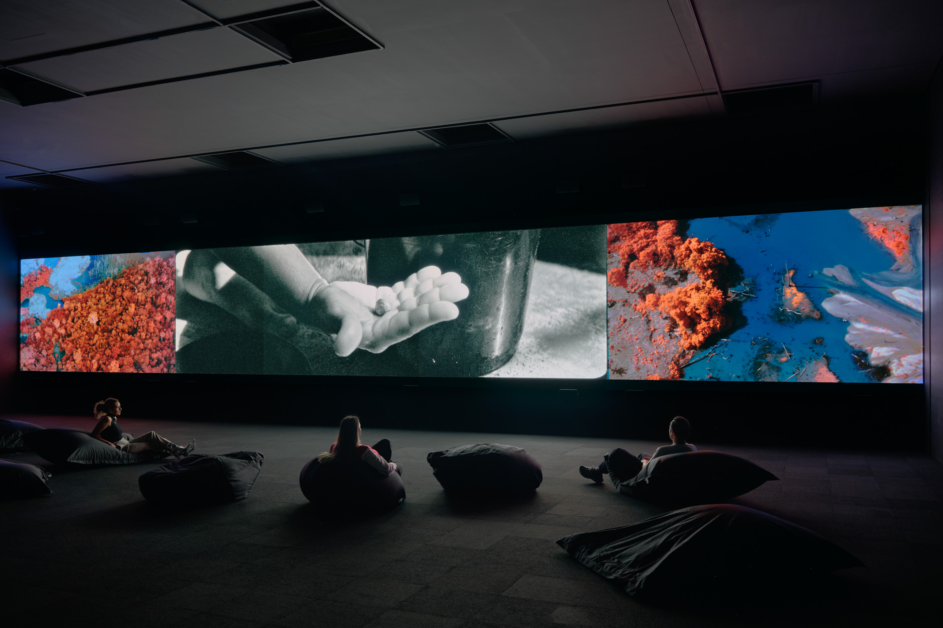 A darkened gallery featuring a panoramic screen with three panels showing different images. People sit on beanbags watching..