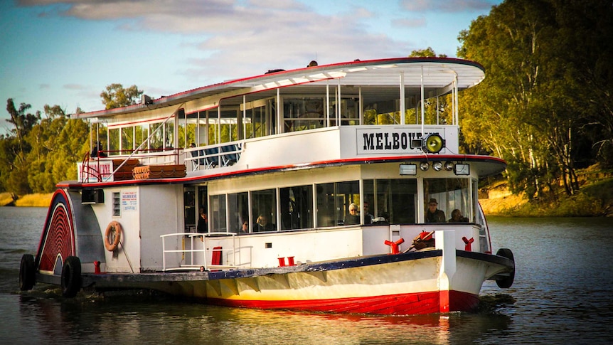 A two-deck white paddle steamer moves up a river with pretty trees in the background.