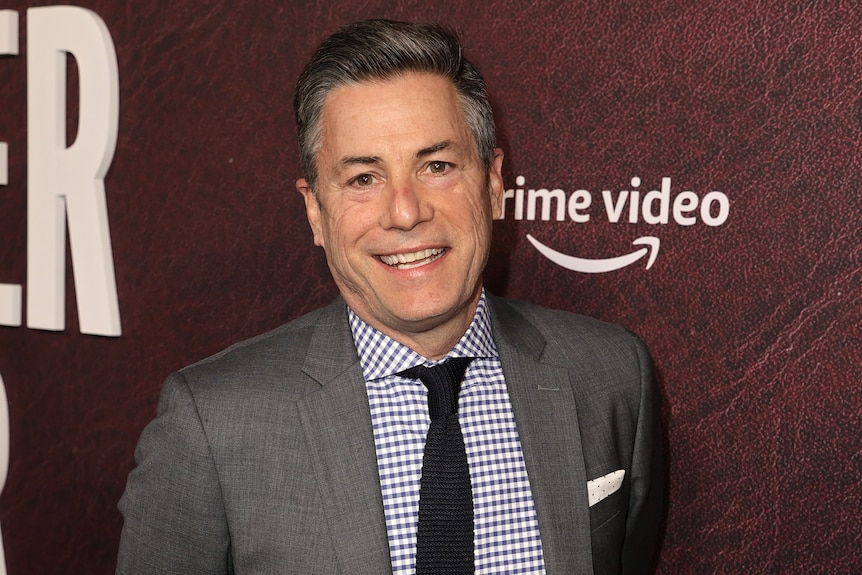 A middle-aged white man wearing a grey suit smiles at a movie premiere