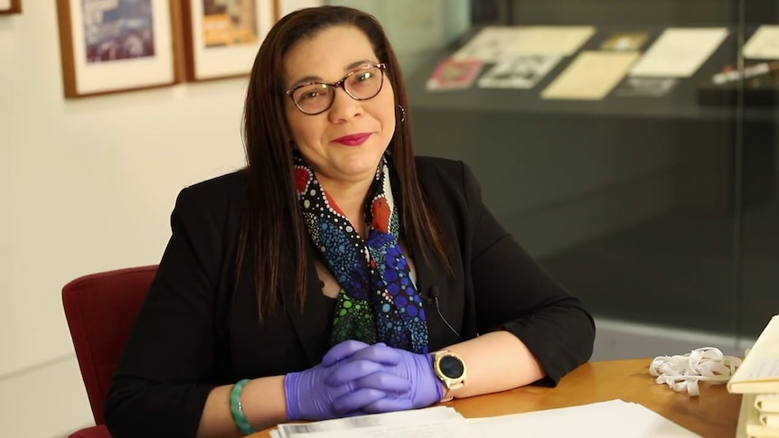 Larrakia woman Danusha Cubillo sits at a desk, wearing gloves, with historical documents in front of her.