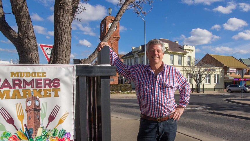 A man standing next to a sign saying Mudgee farmers markets with a streetscape in the background