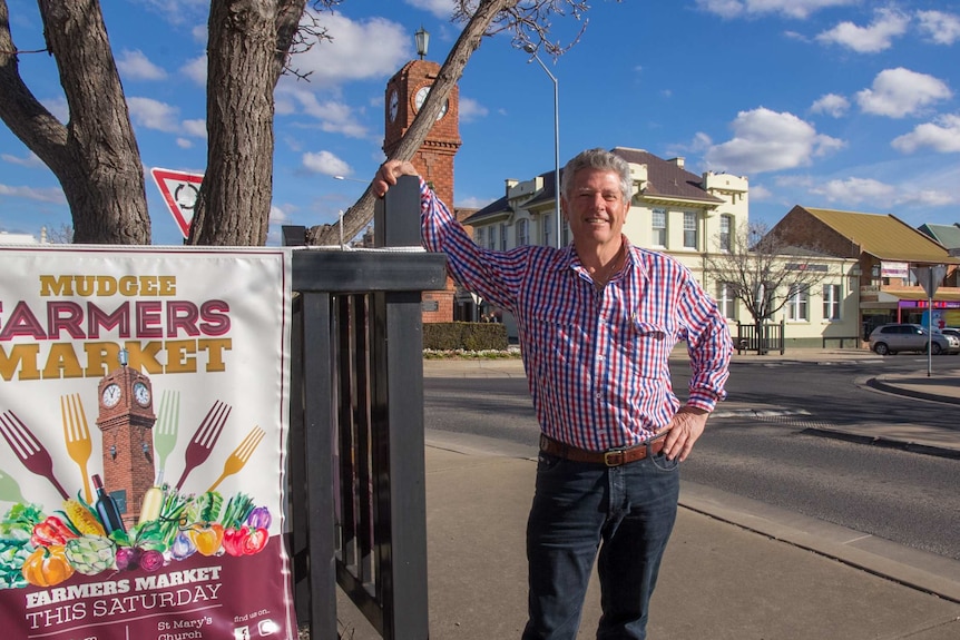 A man standing next to a sign saying Mudgee farmers markets with a streetscape in the background