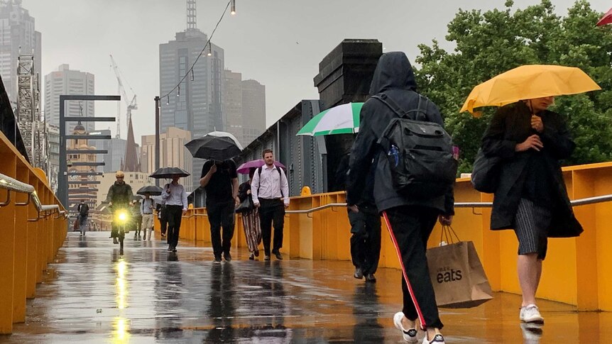 Pedestrians walk over a bridge covered in rain. Many are carrying umbrellas, and one bike has a headlight on.