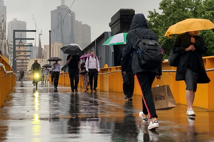 Pedestrians walk over a bridge covered in rain. Many are carrying umbrellas, and one bike has a headlight on.
