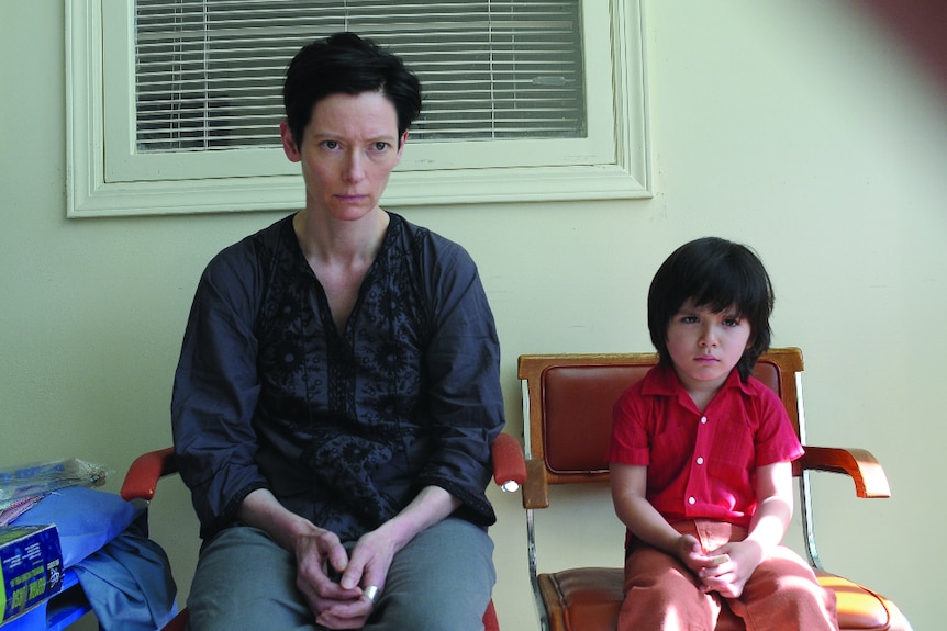 A woman and young child - both dark short hair - with serious expressions sit side by side on waiting room chairs in daytime.