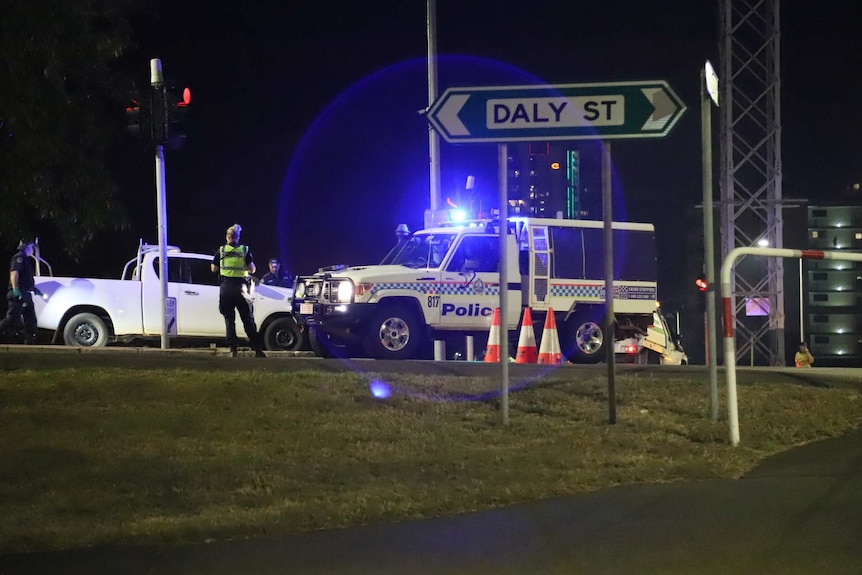 A police vehicle is seen with headlights on. Several officials mill about on road in aftermath of shooting.