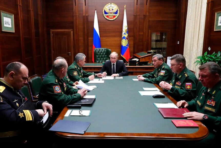 In a dark wood-panelled room, Vladimir Putin sits at the head of a meeting with top military officials in dark green uniforms.