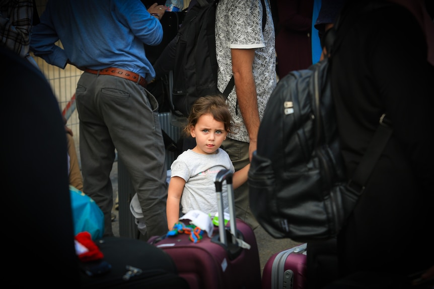 A little girl stands among adults near a suitcase 