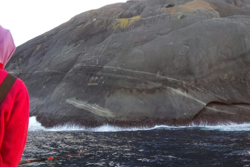 A man fishing from a boat and a man abseiling down a cliff to a wooden ledge