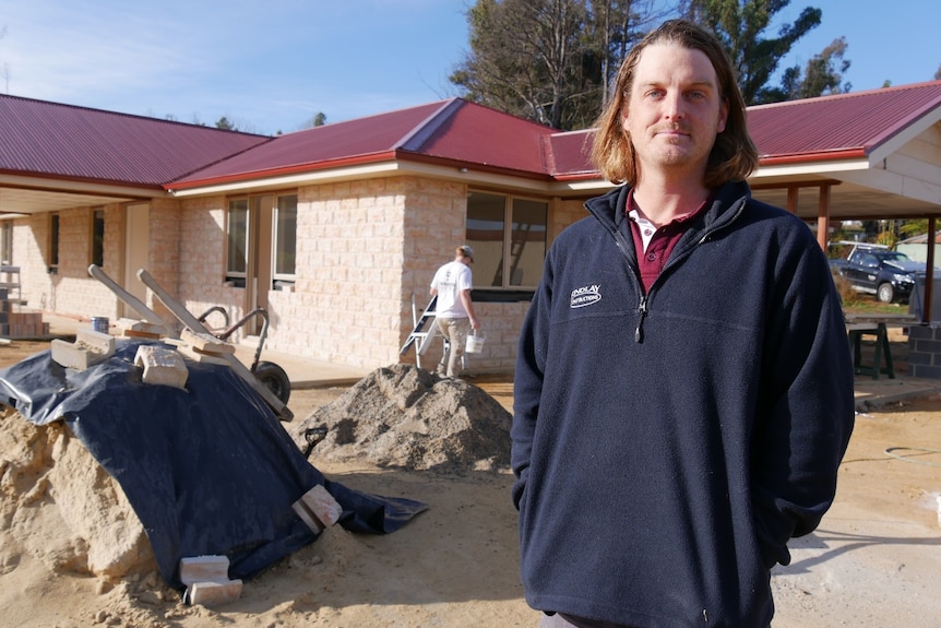 A man with long dark hair stands in front of a house, wearing a jumper.