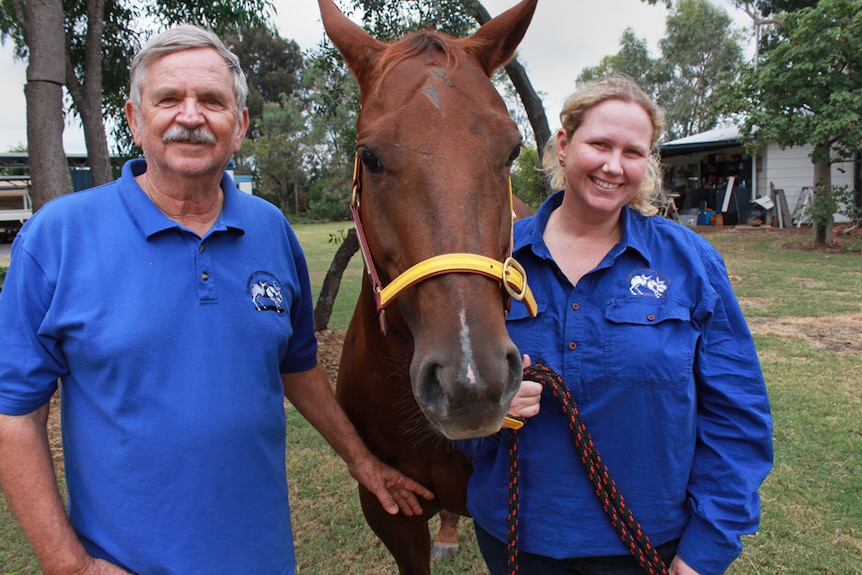 Alan and Tess in blue shirts stand on either side of a brown horse.