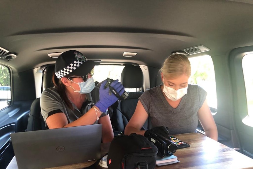 Two police officers sit in a car, one films the other looking at a mobile phone