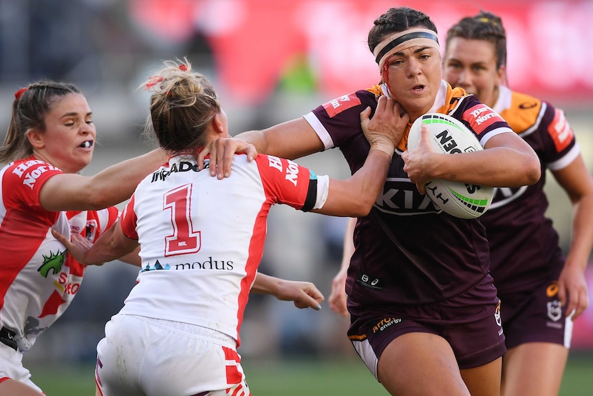 A Brisbane Broncos NRLW player attempts to palm a St George Illawarra opponent while holding the ball with her left hand.