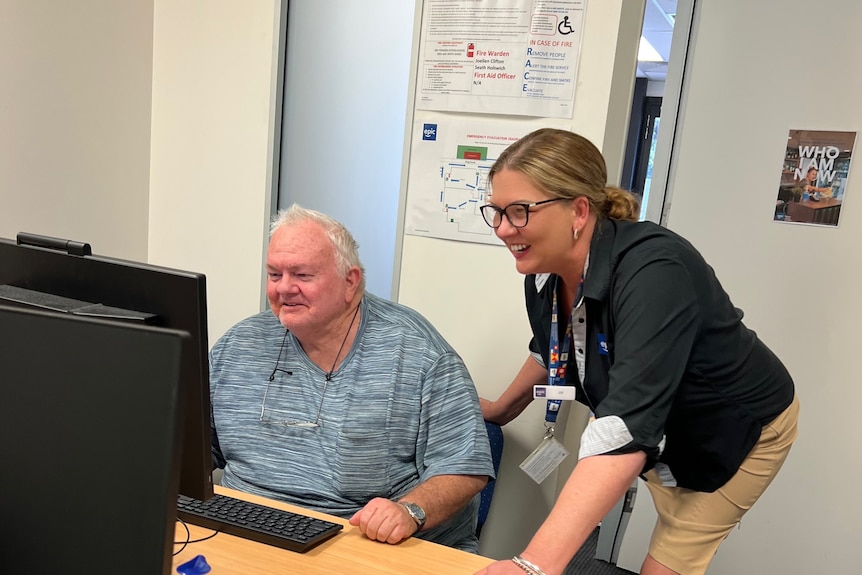 A man sits down looking at a computer with a woman leaning over and looking at it with him.