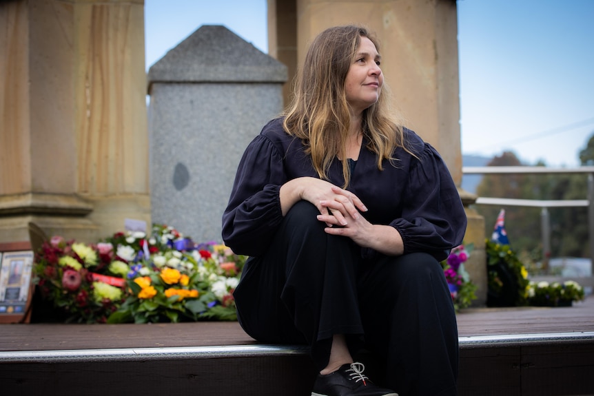 A woman wearing black looks into the distance, while sitting with her back to a memorial with flowers on it. 