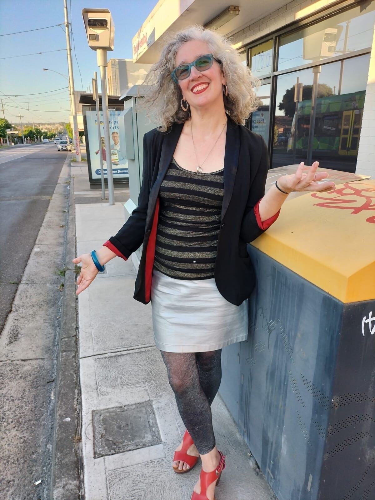 Hilary Harper, with light grey hair, red glasses, shiny top and skirt, stands on a street smiling widely.