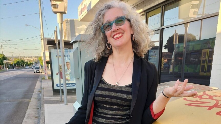 Hilary Harper, with light grey hair, red glasses, shiny top and skirt, stands on a street smiling widely.