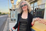 Hilary Harper, with light grey hair, red glasses, shiny top and skirt, stands on a street smiling widely.