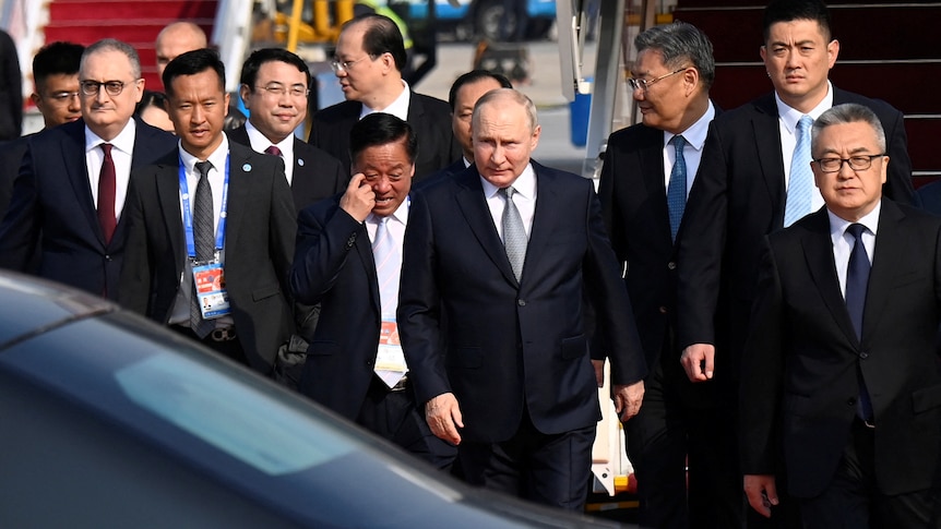 Vladimir Putin emerges from a pack of men in suits, looking proud of himself