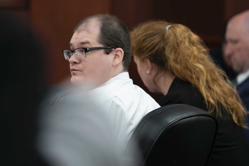 Balding Caucasian man with glasses and white shirt looks over his shoulder while sitting in the court room.