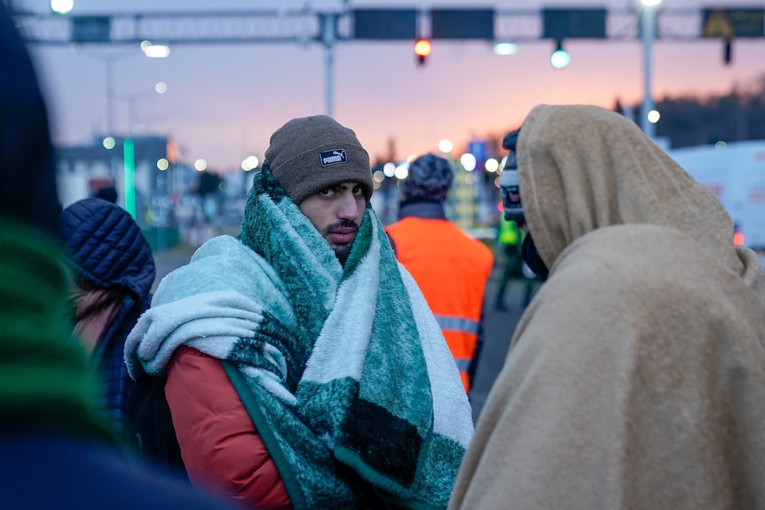 A man wearing a beanie, winter coat and draped in a blanket, stands in a crowd of people walking towards a border checkpoint