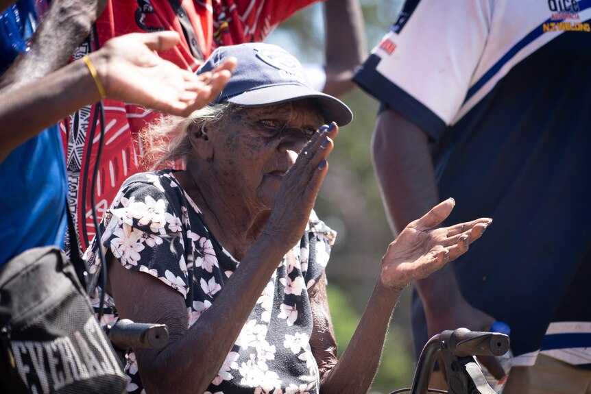An elderly Indigenous woman claps on the sidelines of a football field.