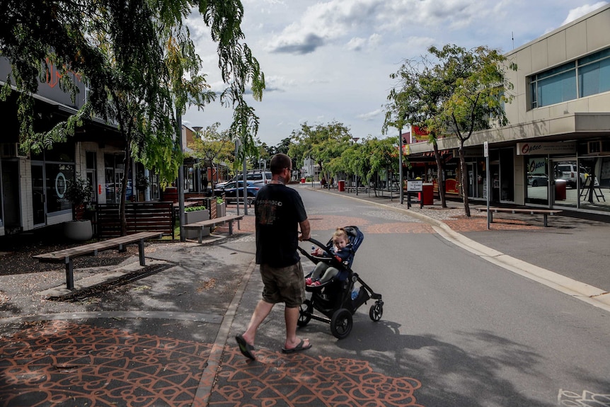 Man wearing black t-shirt with tattoos with young daughter in pram across street with shopping strip other side