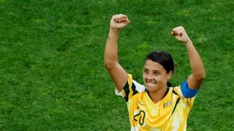 A woman wearing a Matildas soccer team jersey holds her fists above her head.
