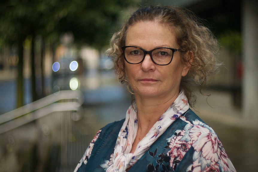 A woman wearing glasses and a floral outfit looks straight ahead without smiling.