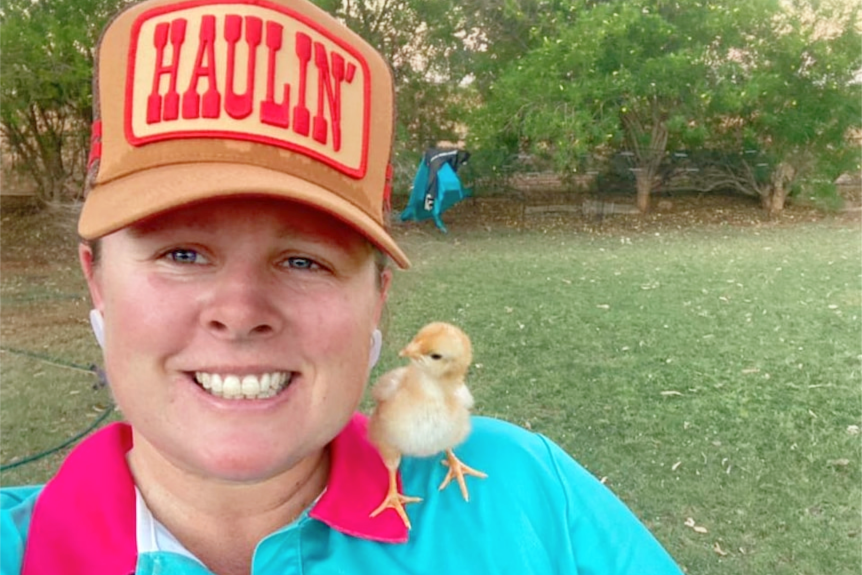 A woman with an orange hat and a blue shirt smiles, while a fluffy yellow chick sits on her shoulder.