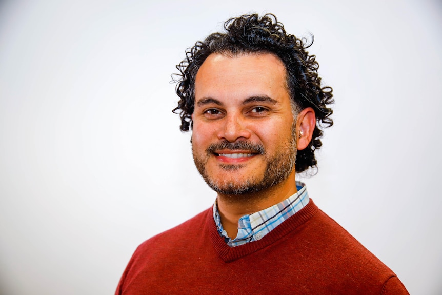 A man with curly hair wearing a red sweater smiles at the camera in front of a white background