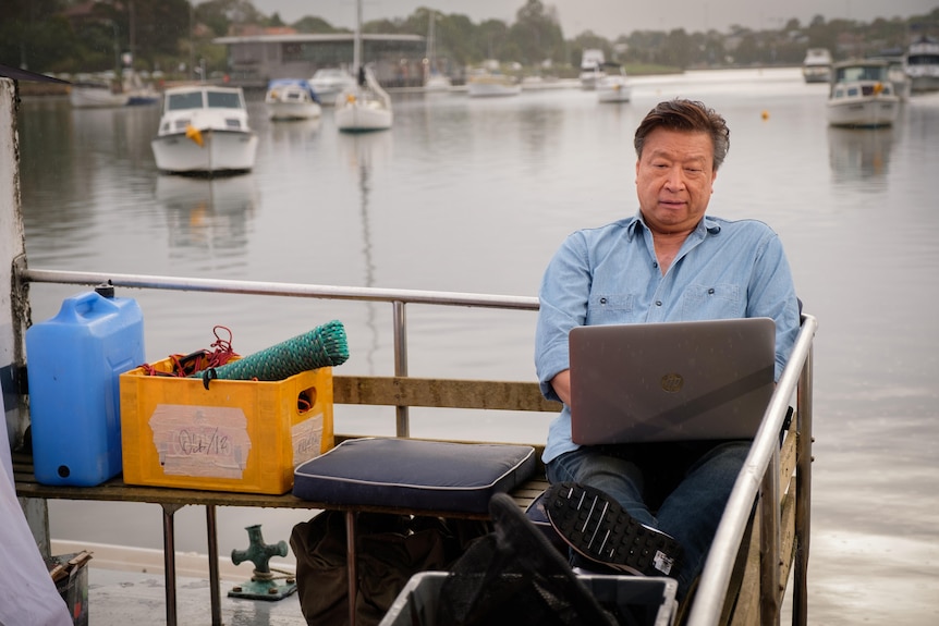 Tzi Ma sits on a wharf with his feet up as he peers intently at a laptop, boats seen on a harbour behind him.